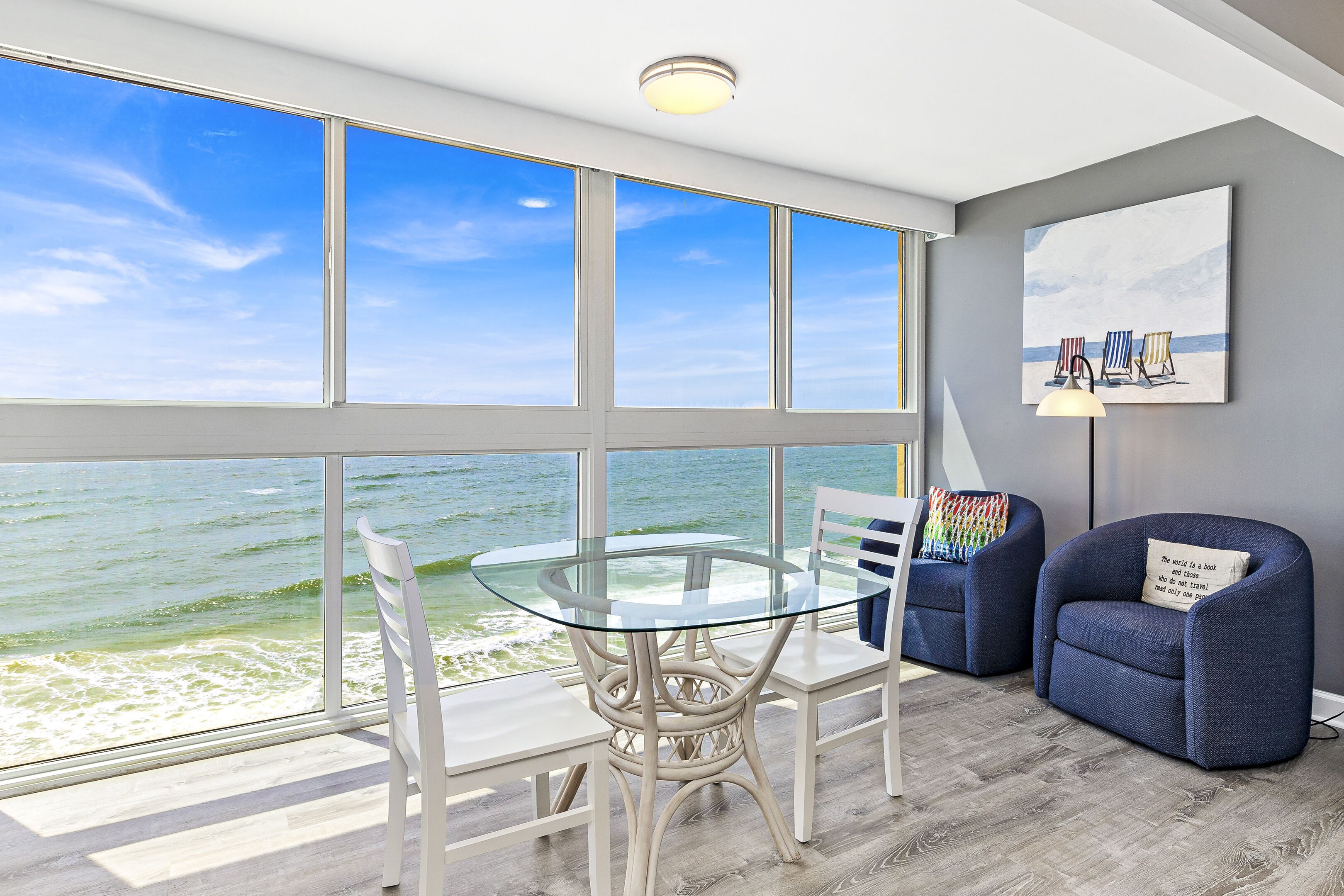 A room with a glass table and chairs, a blue armchair, and a painting of beach chairs on the wall.