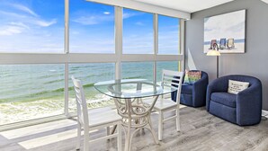 Une pièce avec une table et des chaises en verre, un fauteuil bleu et un tableau représentant des chaises de plage accroché au mur.