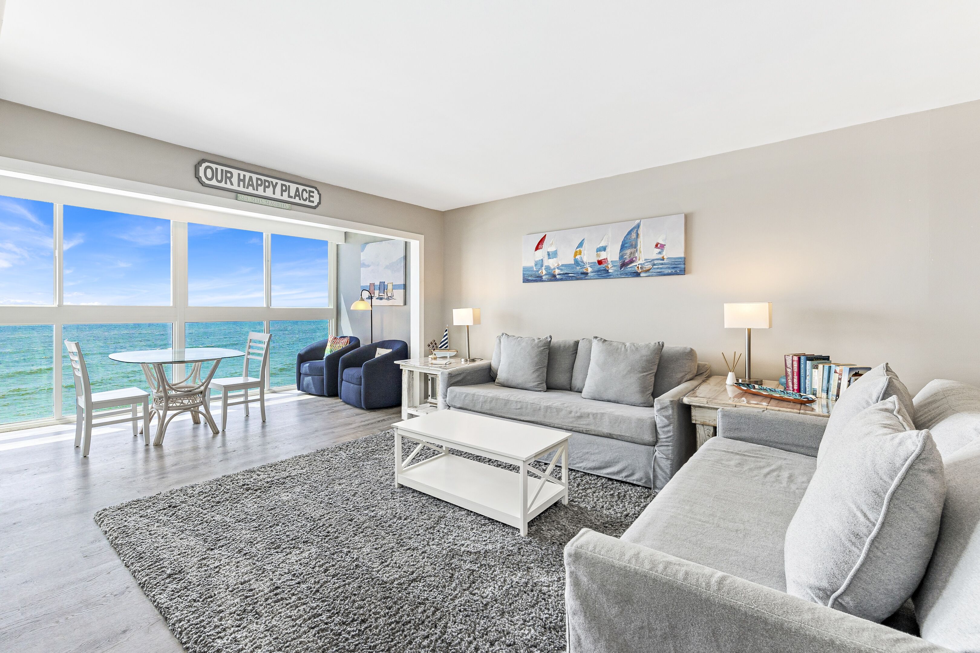 A modern living room with a large window overlooking the sea, a cozy seating area with a sofa and armchairs, a small round table, and a coffee table.
