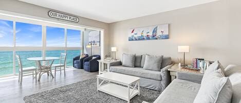A modern living room with a large window overlooking the sea, a cozy seating area with a sofa and armchairs, a small round table, and a coffee table.