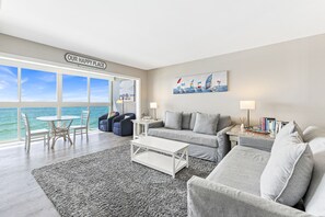 A modern living room with a large window overlooking the sea, a cozy seating area with a sofa and armchairs, a small round table, and a coffee table.