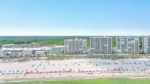 Une vue sur la plage, avec de nombreux immeubles de grande hauteur, une plage de sable parsemée de parasols et une zone herbeuse.
