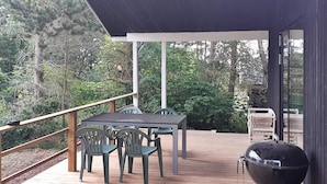 Une terrasse extérieure couverte, équipée d’une table et de chaises, d’un barbecue et offrant une vue sur la forêt environnante.