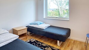 A small, single-bed room with a window, a bedside table, and a chair.