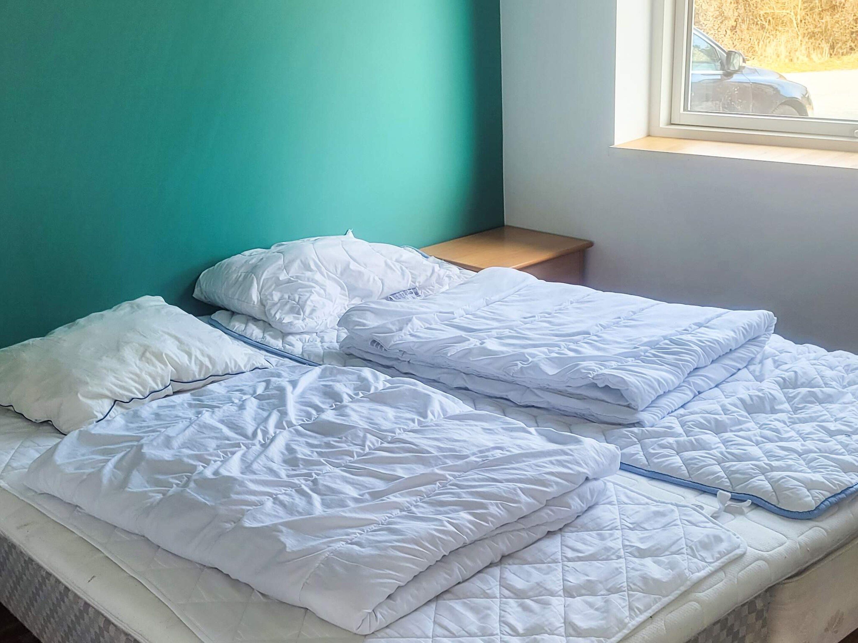 A bed with white bedding and pillows, a wooden nightstand, and a window with a view of a car and trees.