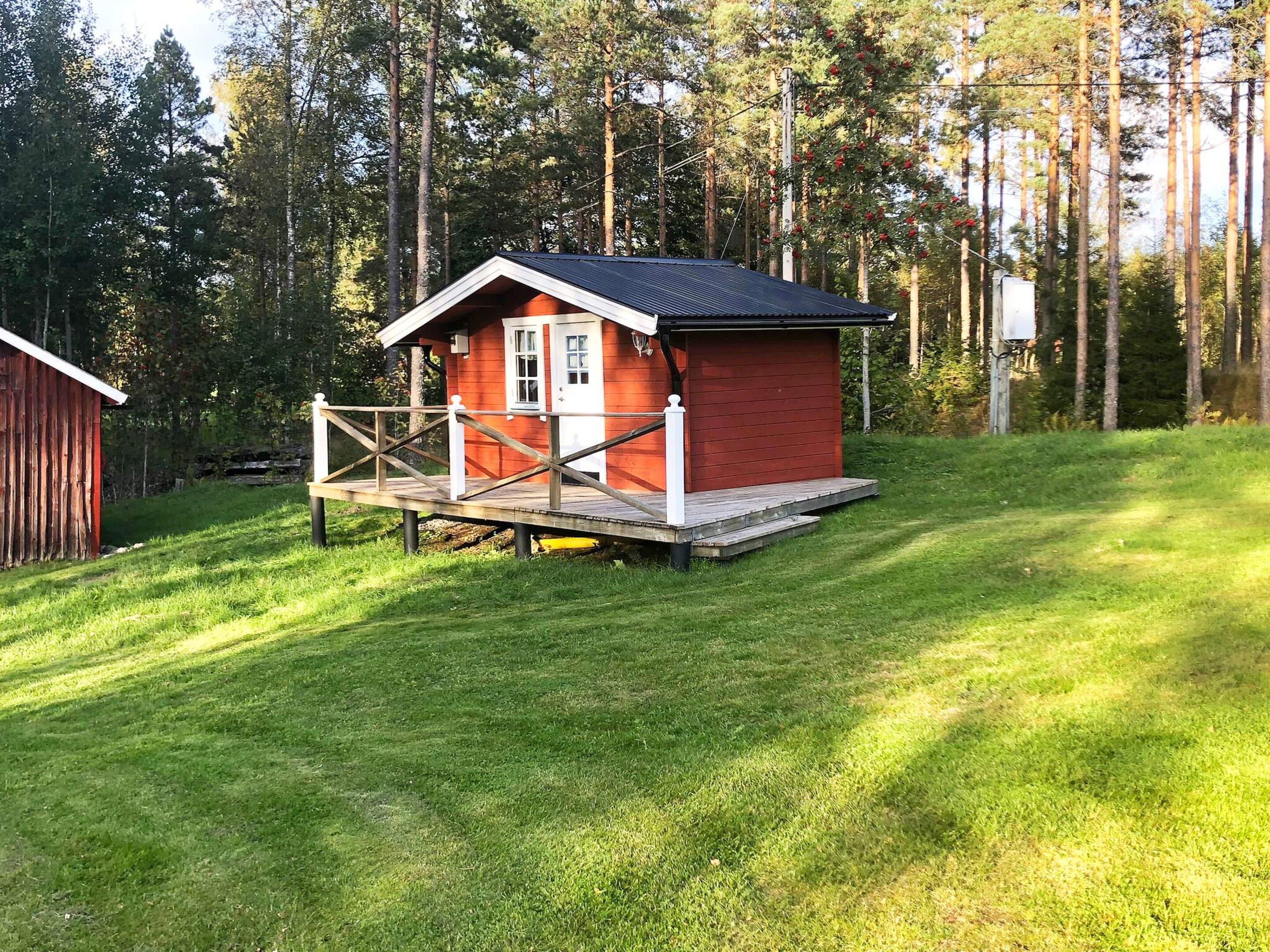 A small red cabin with a white porch and a wooden deck, surrounded by a grassy area and tall trees.