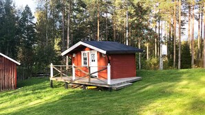 Une petite cabane rouge avec un porche blanc et une terrasse en bois, entourée d’une pelouse et de grands arbres.