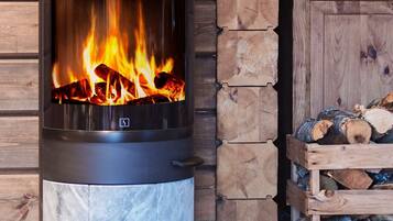 A wood-burning stove with a visible fire, set against a wooden wall with a log storage area nearby.