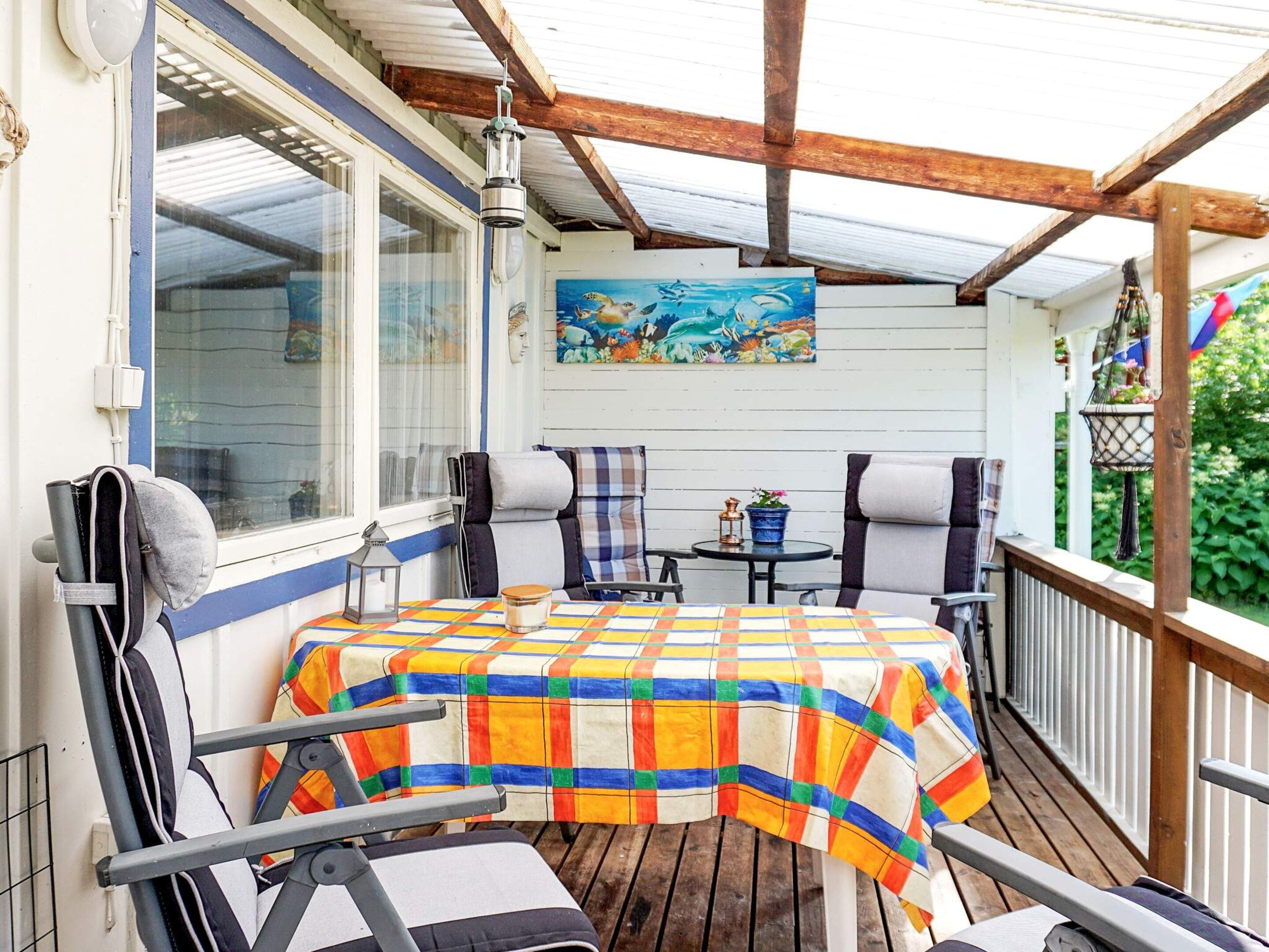 A covered outdoor seating area with a table and chairs, a painting on the wall, and a window with curtains.