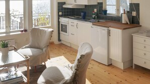 A modern kitchen with white cabinets, a stove, and a sink. There is a chair covered with a cloth, a small table with a plant, and a view of a lake through large windows. - 4 Person Holiday Home in Farentuna (Faerentuna)