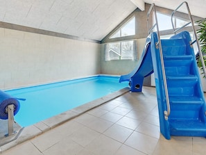 An indoor swimming pool with a blue slide and a ladder, surrounded by tiled walls and windows.