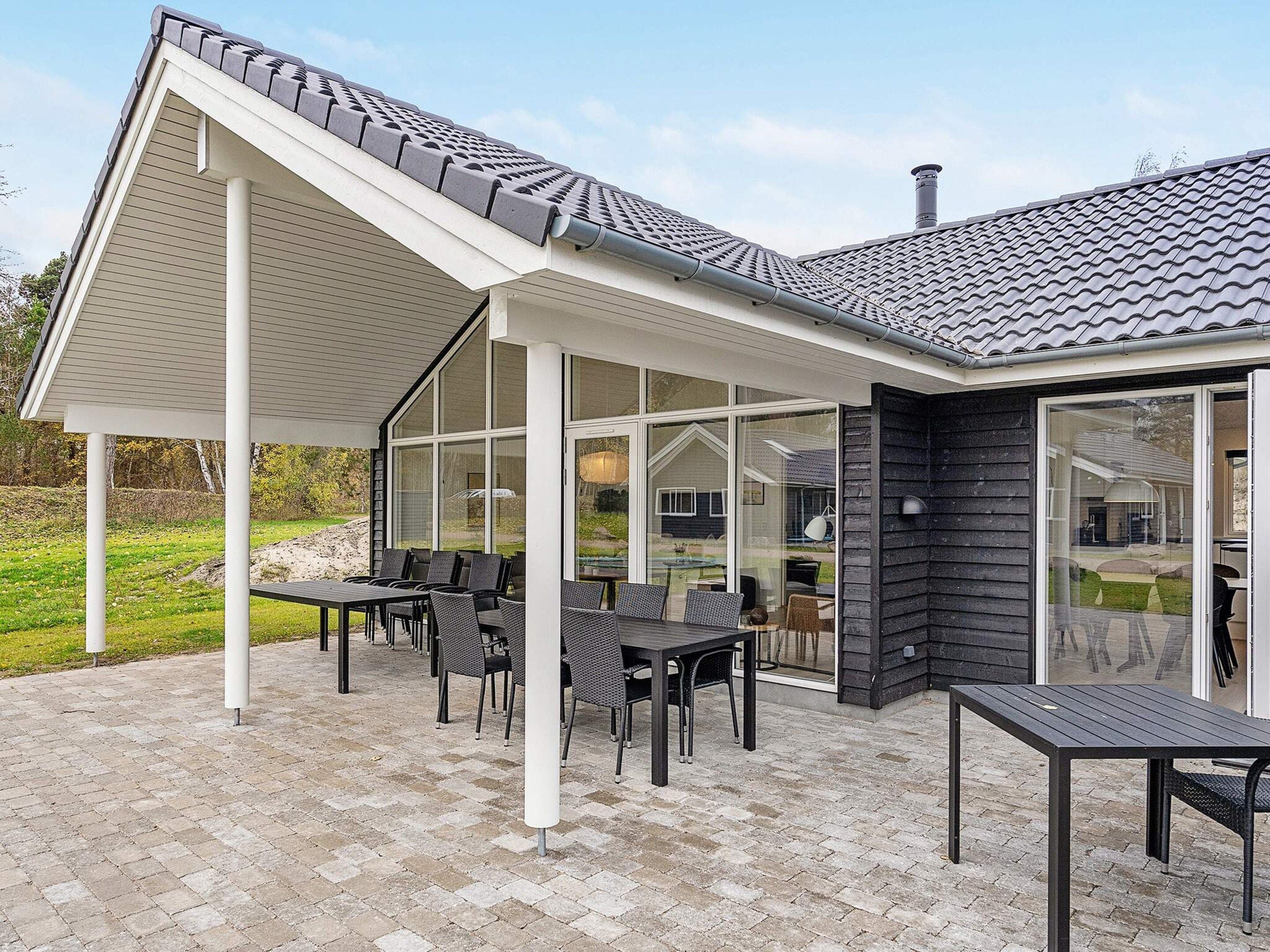 Un espace extérieur aménagé, abrité et doté de tables et de chaises, une structure en forme de pergola et une vue sur la maison à travers de grandes fenêtres.
