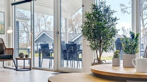 A room with a large sliding glass door leading to an outdoor patio area. Inside, there is a round wooden table with a bench, a wicker chair, and a small side table with decorative items. - 5 Star Holiday Home in Vaeggerlose (Vaeggerlose)
