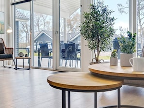 A room with a large sliding glass door leading to an outdoor patio area. Inside, there is a round wooden table with a bench, a wicker chair, and a small side table with decorative items. - 5 Star Holiday Home in Vaeggerlose (Vaeggerlose)
