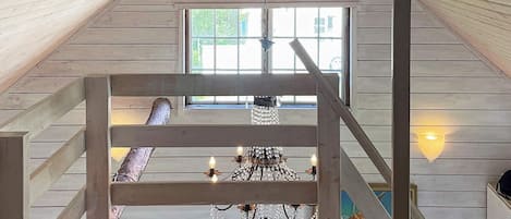 A wooden interior with a staircase leading to a balcony, a chandelier, and a window with a view.