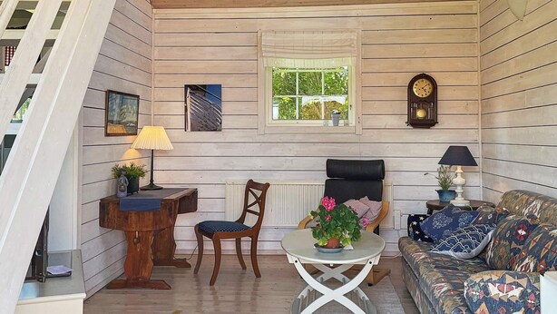 A cozy room with a wooden ceiling, a dining table with chairs, a sofa, a clock, and a window with a view of greenery. - 4 Star Holiday Home in Ellos (Ellos)