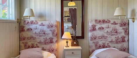 A room with two beds, a nightstand with a lamp, a mirror, and patterned wallpaper.