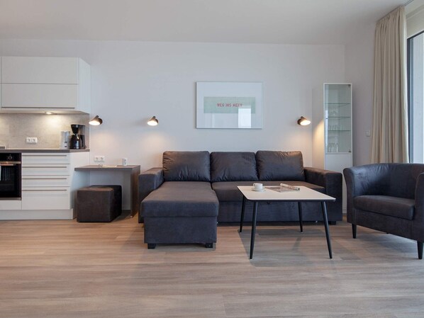 Uma sala moderna com um sofá cinzento, uma mesa de centro e um espaço de refeições, com uma cozinha ao fundo.