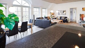 Een ruime woonkamer met een eettafel, stoelen, banken en een salontafel. Grote ramen laten veel natuurlijk licht binnen.