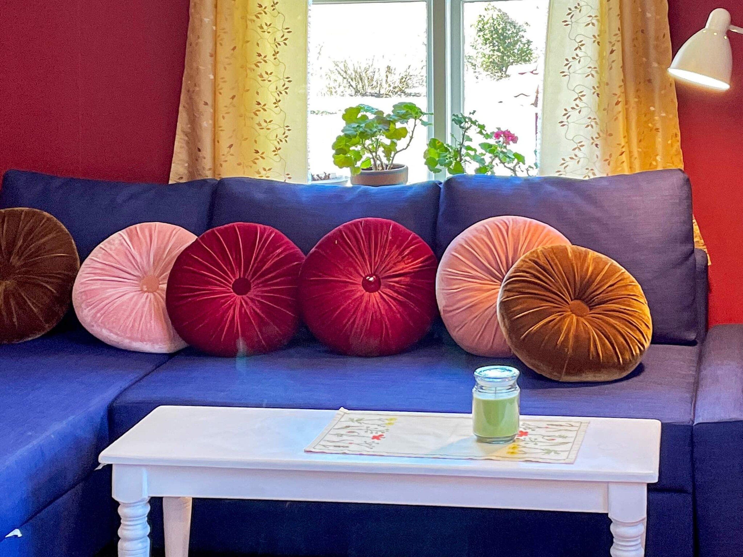 Um sofá azul com cinco almofadas redondas em várias cores, uma mesa branca com um pote de vidro, e uma janela com cortinas e plantas.