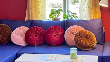 A blue sofa with five round cushions in various colors, a small white table with a glass jar, and a window with curtains and a potted plant.
