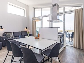 A modern living room with a white dining table, dark chairs, a sofa, and a large window with curtains. - 5 Star Holiday Home in Wendtorf (Lubeck)