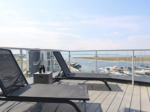Uma varanda com duas poltronas de descanso, uma mesa pequena e vista para um porto com barcos.