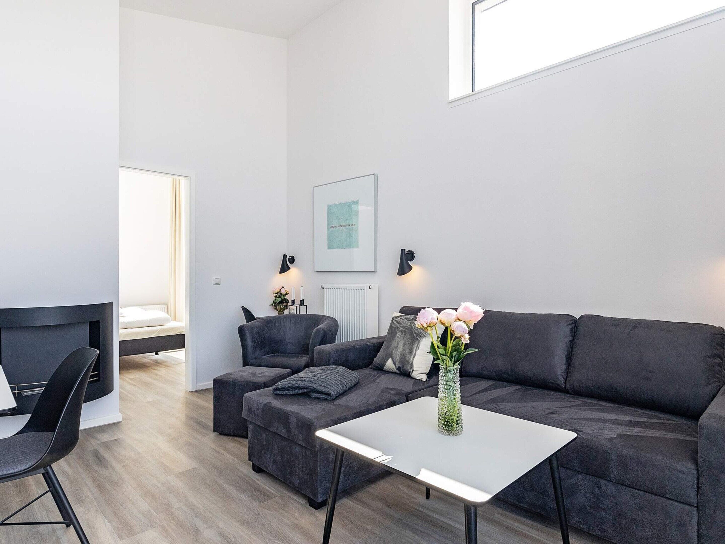 Ein modernes Wohnzimmer mit einer Couch, einem Sessel, einem Hocker, einem kleinen Tisch und einer Vase mit Blumen.