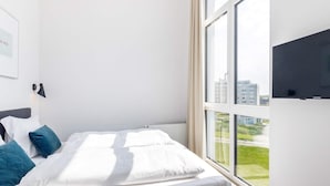 A modern hotel room with a large window, a bed with white bedding and blue pillows, a mounted flat-screen TV, and a wall-mounted light fixture. - 5 Star Holiday Home in Wendtorf (Lubeck)
