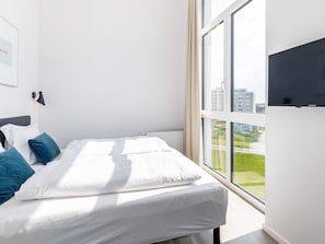 Quarto de hotel moderno com uma janela ampla, cama com roupa de cama branca e almofadas azuis, televisão de tela plana montada e luminária fixa na parede.