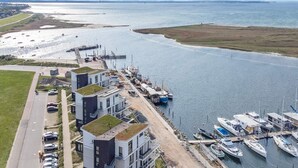 A coastal marina with boats docked, a road with parked cars, and modern buildings with green roofs.