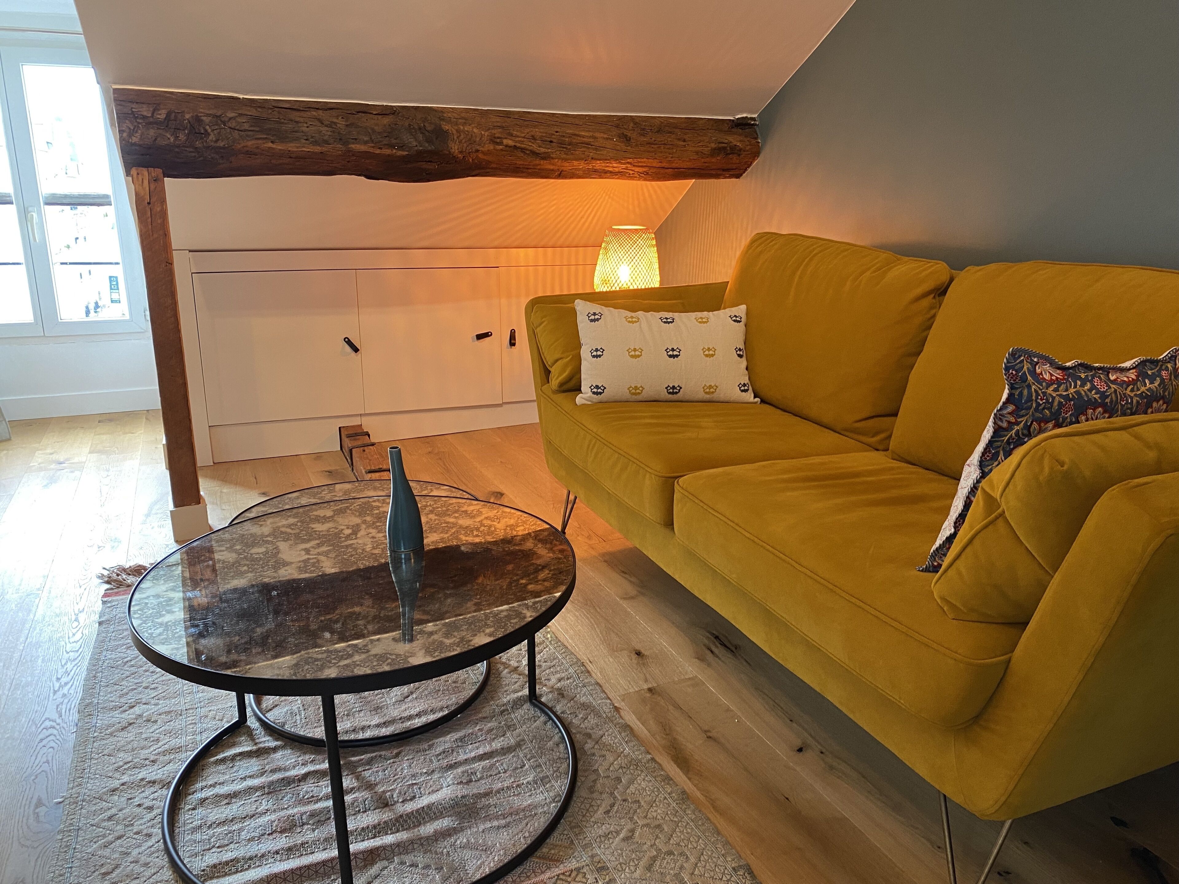 A yellow sofa with patterned pillows, a glass coffee table with a vase, and a wooden beam ceiling.
