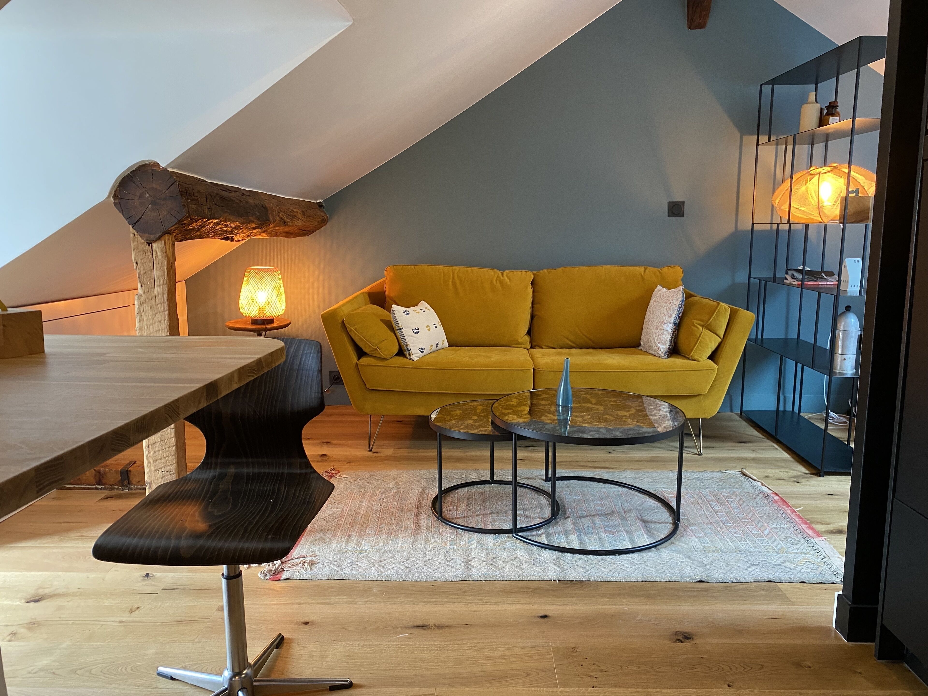 A cozy loft bedroom with a wooden dining table, a yellow sofa, a small coffee table, and a bookshelf.