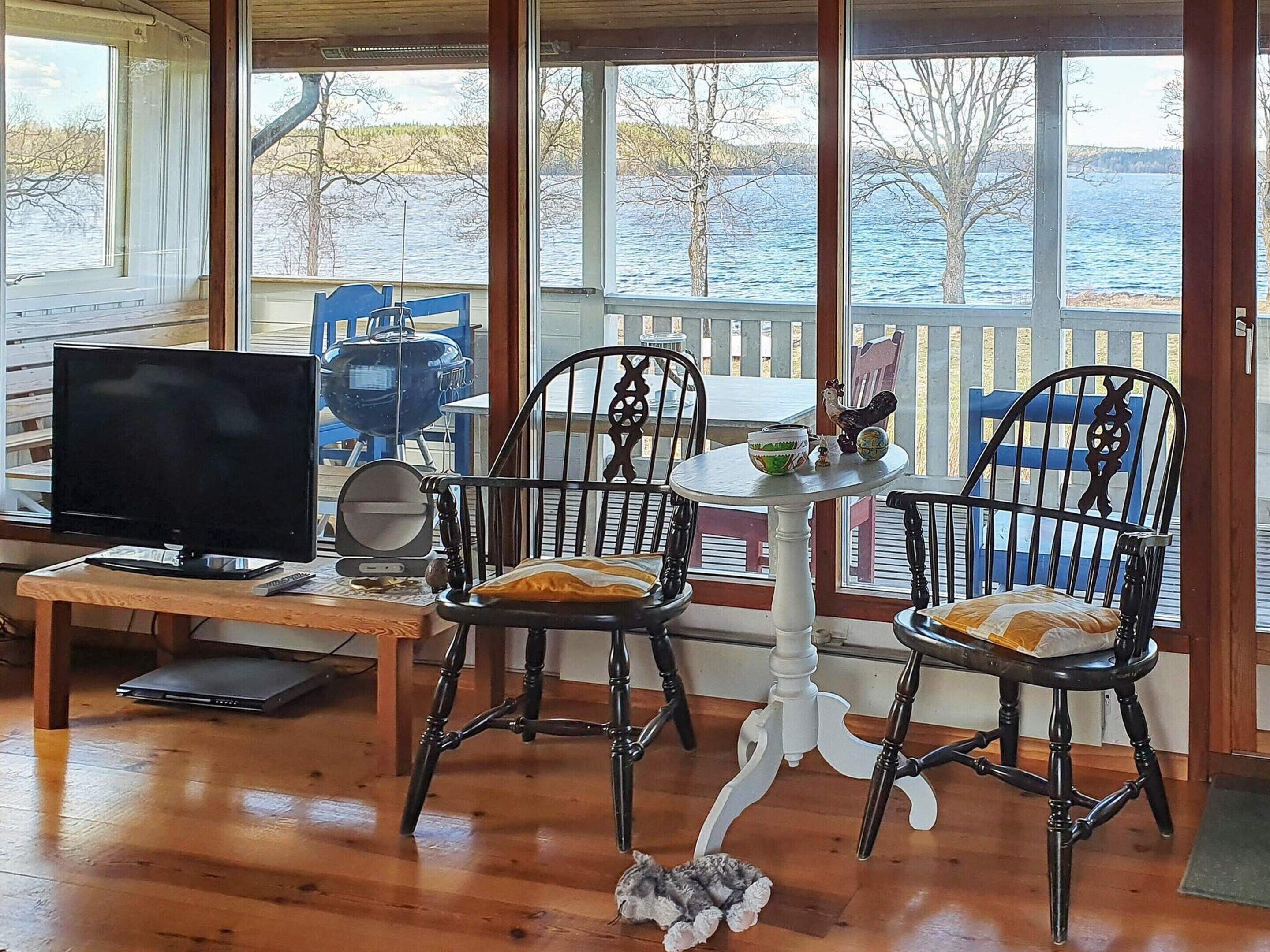 Quarto com piso de madeira, televisão, mesa e cadeiras, e vista para um lago através de grandes janelas.