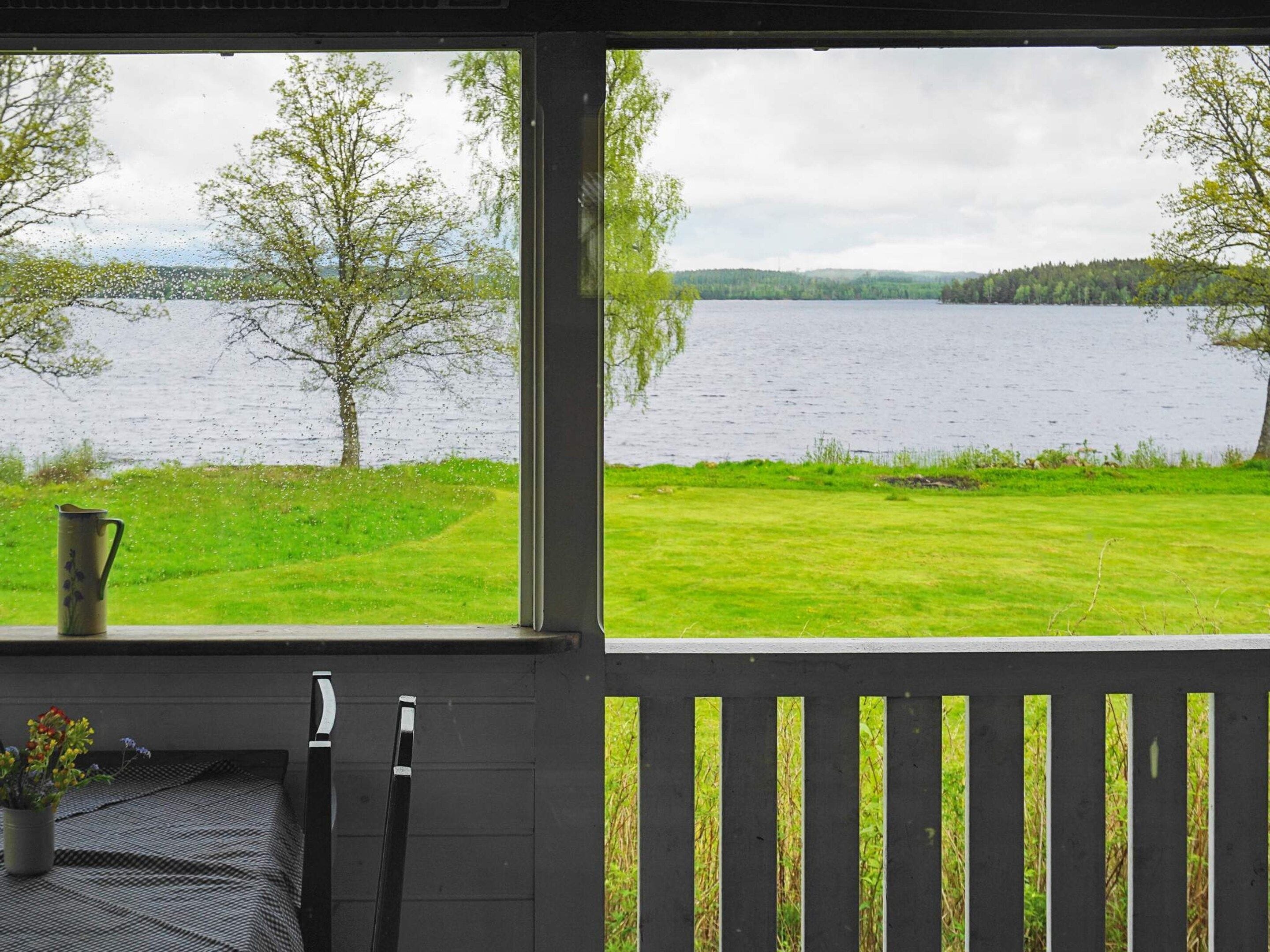 A room with a view of a lake, a wooden deck, and a table with a pitcher and flowers.