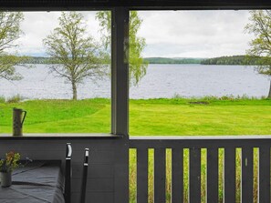 A room with a view of a lake, a wooden deck, and a table with a pitcher and flowers. - 6 Person Holiday Home in Holsljunga (Holsljunga)