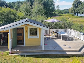 A small yellow cabin with a deck, a table, and chairs, surrounded by greenery and a lake. - 6 Person Holiday Home in Holsljunga (Holsljunga)