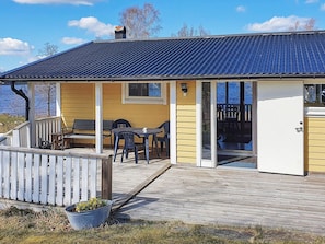 A small, single-story building with a covered patio area, a wooden deck, and a view of the water. - 6 Person Holiday Home in Holsljunga (Holsljunga)