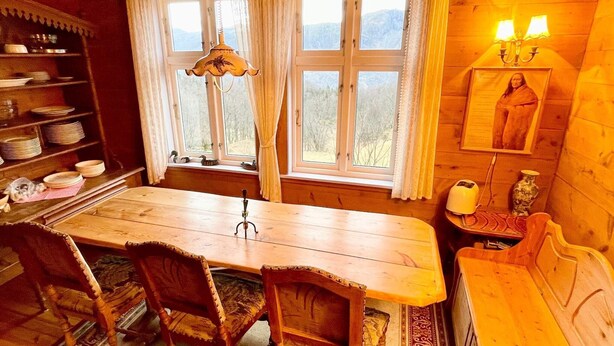 A cozy wooden dining area with a table, chairs, and a bench. There are windows with curtains, a hanging lamp, and a shelf with decorative items. - 8 Person Holiday Home in Akra-by Traum (Kvinnherad)