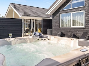 A hot tub with a glass enclosure, a dining area with chairs and a table, and a house with a dark exterior and a tiled roof.