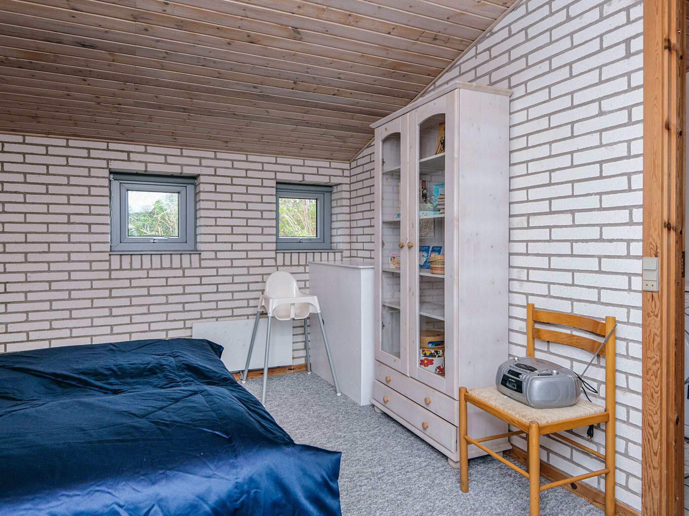 A bedroom with a wooden ceiling, white brick walls, a bed with a blue cover, a white wardrobe, and a wooden chair.