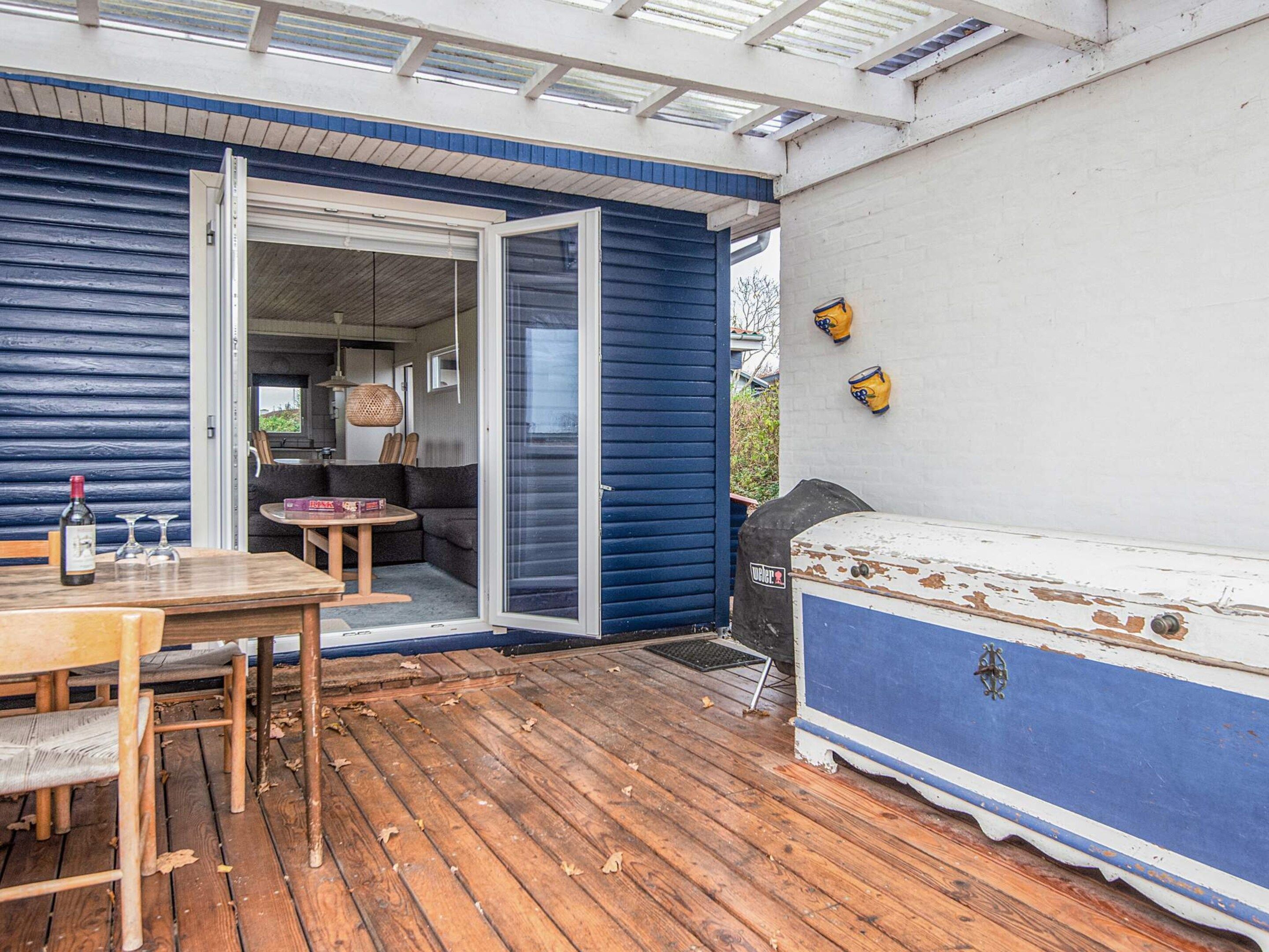 A wooden deck with a table and chairs, a blue and white outdoor seating area, and a glass door leading to an interior space with a sofa and a table.