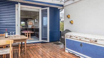 A wooden deck with a table and chairs, a blue and white outdoor seating area, and a glass door leading to an interior space with a sofa and a table.