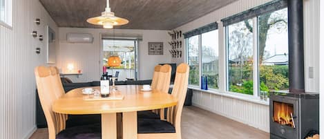 A dining area with a wooden table and chairs, a hanging light fixture, a fireplace, and large windows with a view of the outdoors.