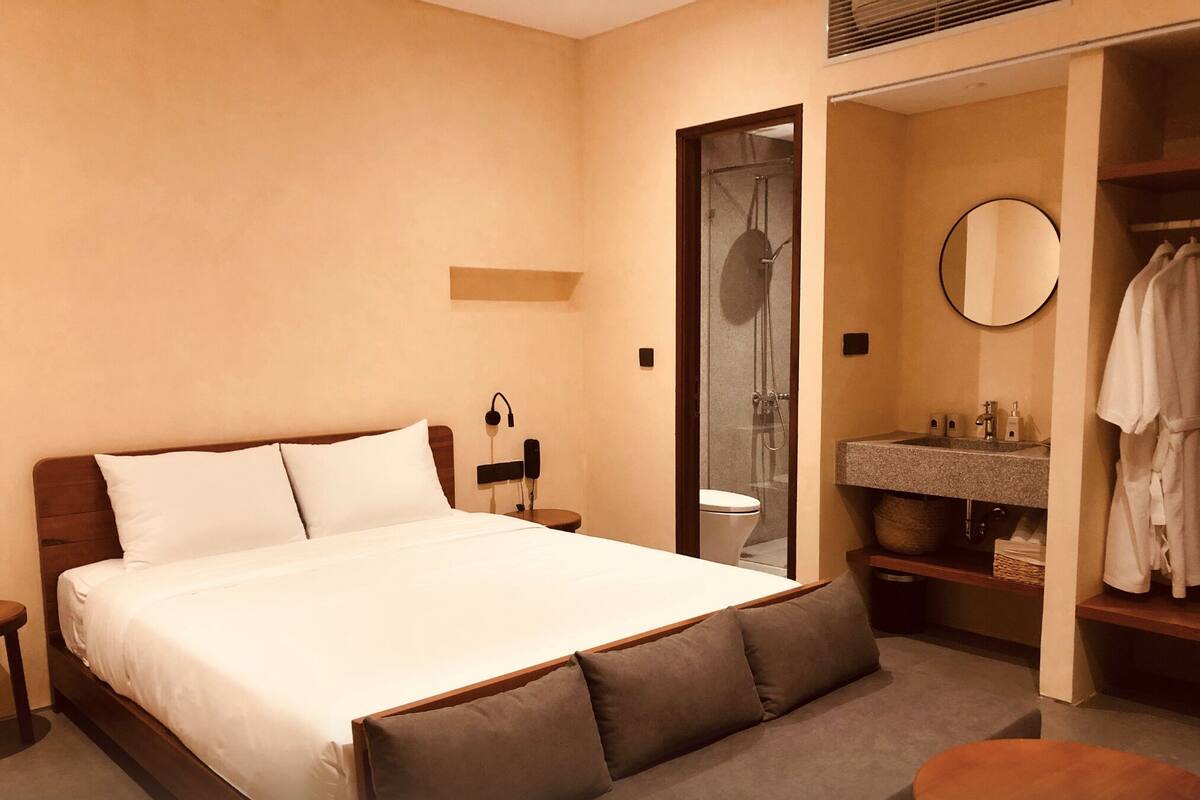 A hotel room with a large bed, a bedside table, a mirror, a bathroom with a sink and a robe hanging.