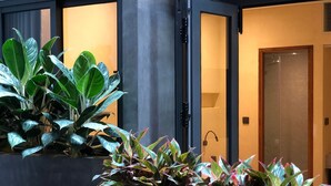 A modern hotel room with a large potted plant in the foreground, a sliding door to the right, and a plain wall with a door at the end. - Shelter Stay Da Nang (Da Nang)