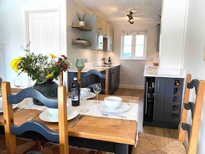 Un comedor con una mesa de madera dispuesta para dos personas, un jarrón de flores y una cocina al fondo.