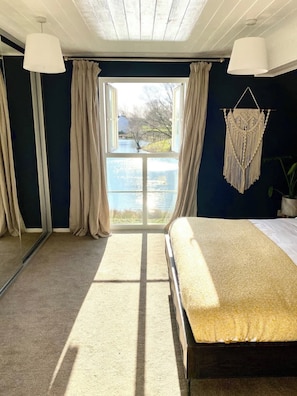Een slaapkamer met een groot bed, uitzicht op een meer en een wanddecoratie van macramé.