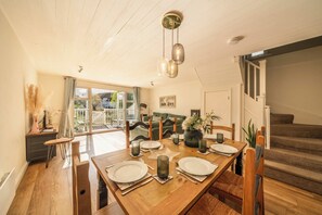 A dining area with a wooden table set for four, a staircase to the right, and a view of the outside through a glass door. - South Facing Cotswolds Water Park Lake House. (Cirencester)