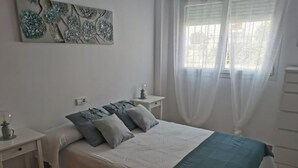 Un dormitorio con cama, mesitas de noche, lámpara y ventana con cortinas translúcidas.